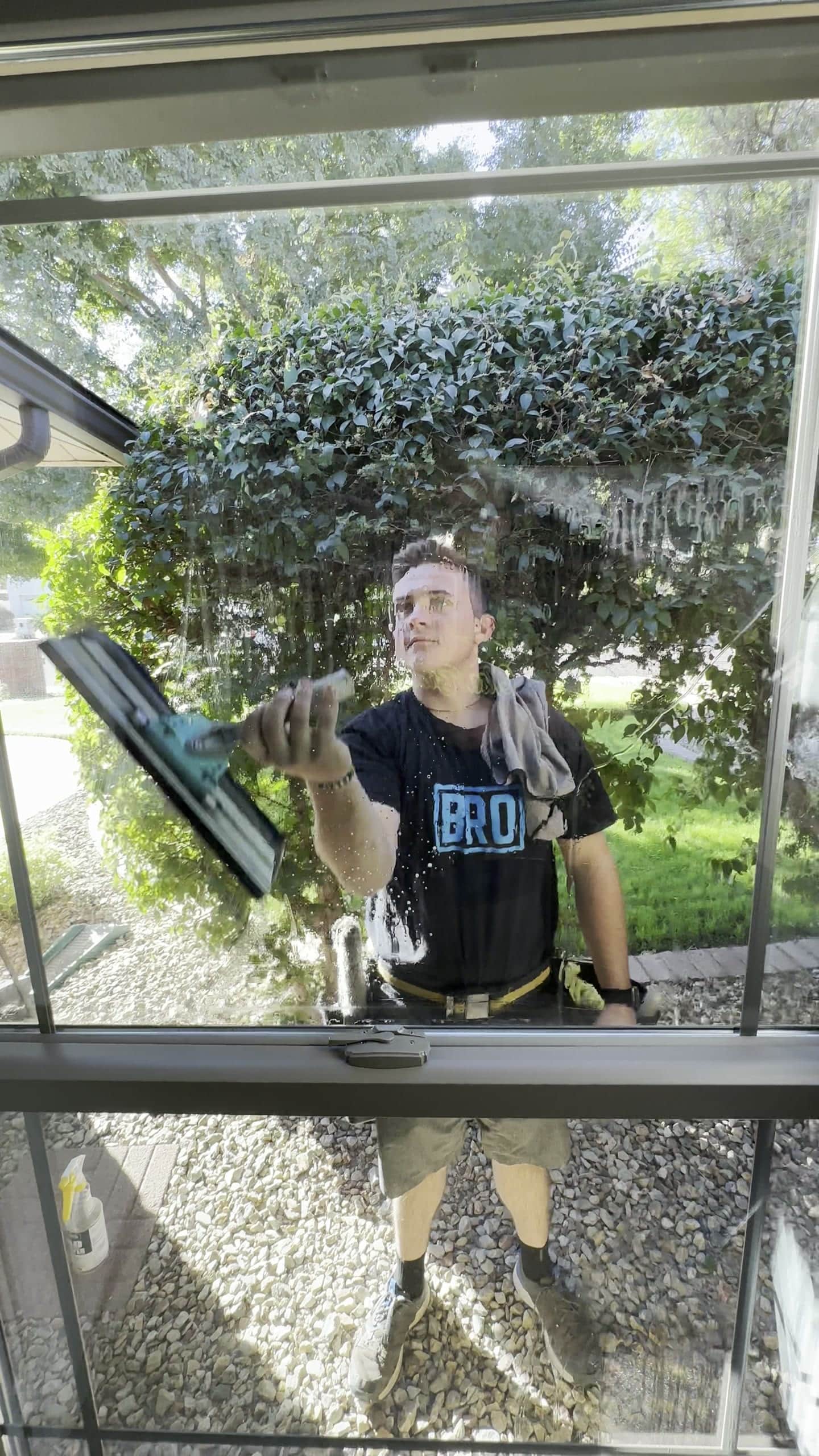 Olsen Brothers window cleaner in a BRO shirt squeegeeing a homeowner's window, seen from inside the house, East Valley, Arizona