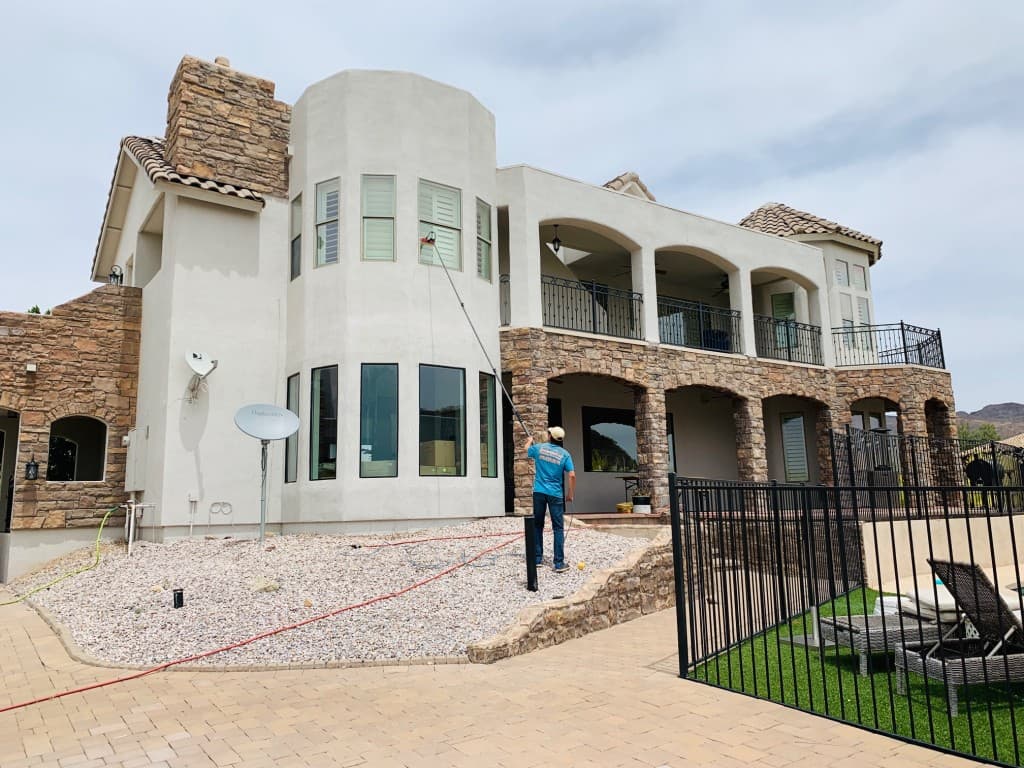 Technician using a water-fed pole to clean second-story windows on a large stucco and stone home — professional residential window cleaning, Arizona